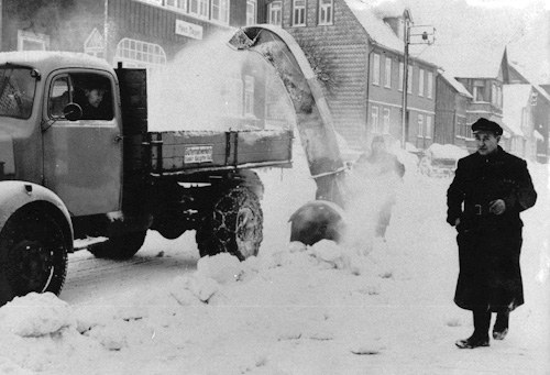 Lastwagen mit aufgebauter Schneeschleuder auf einer tief verschneiten Ortsstraße; der Schnee fliegt im Bogen aus dem Auswurf, rechts geht ein Mann im Mantel vorbei.
